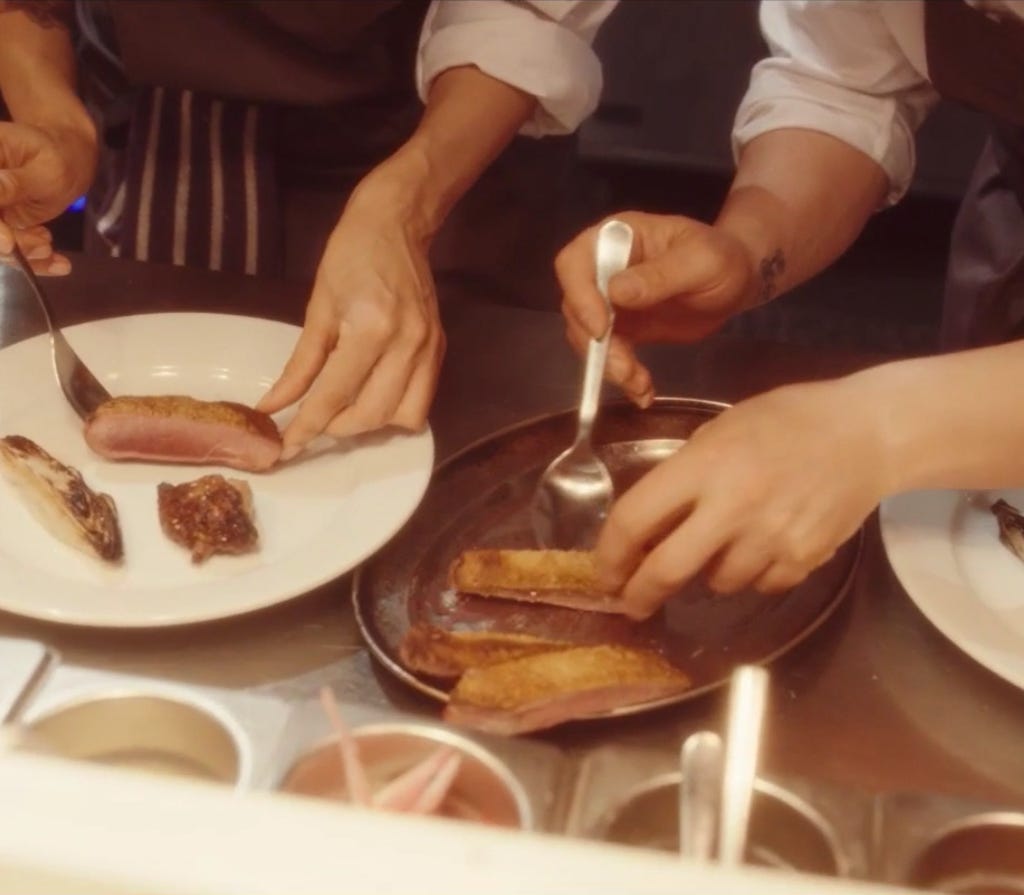 A still from 'Boiling point', two pairs of white hands plate up meat at a pass, under warm light. 