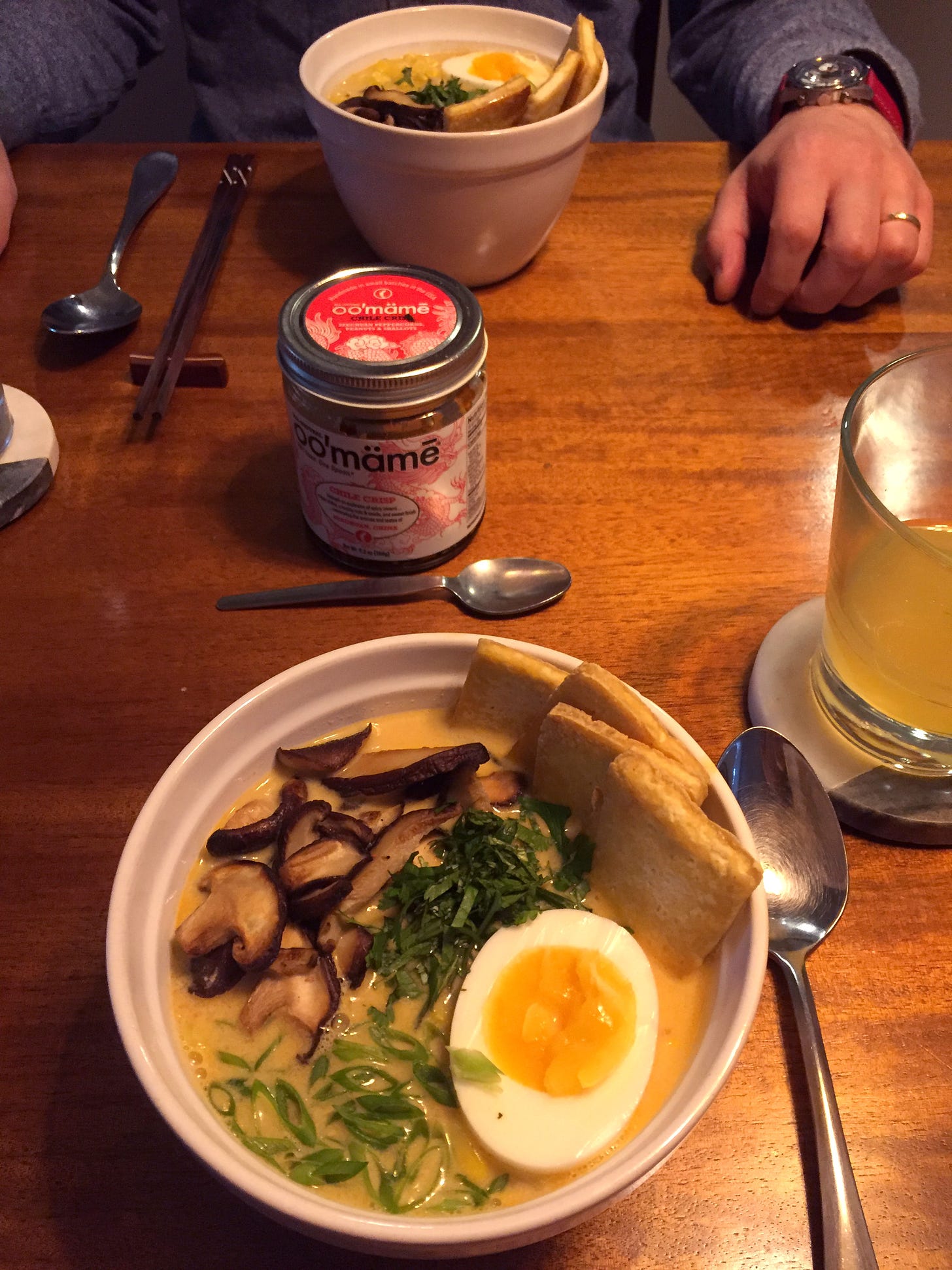 Two bowls of ramen across from each other on the table, with a jar of chili crisp in the middle. In each bowl are slices of tofu, fried shiitake mushrooms, green onions, corn, and half a soft-boiled egg.