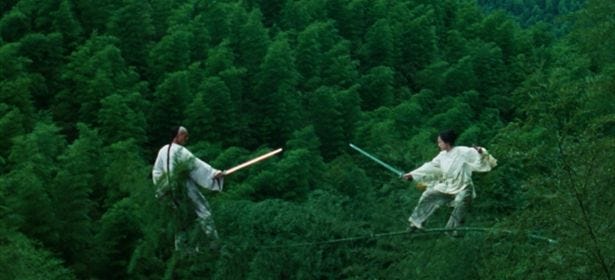 Print de cena do filme "O tigre e o dragão". Na imagem, dois personagens vestidos de branco se equilibram sobre o fino galho de um bambu, em meio à um gigantesco bambuzal. As personagens portam espadas e as apontam em direção um ao outro, prestes a atacar. Ao fundo, tudo verde devido ao bambuzal.