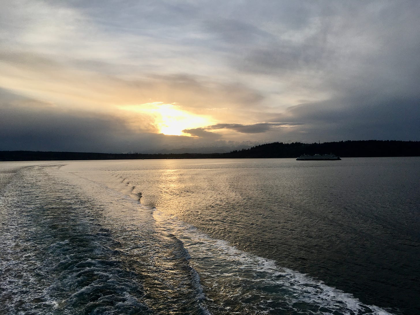 setting sun behind clouds and ferry boat in distance. picture is taken on other ferry boat and so, water flow reveals path of ferry boat.
