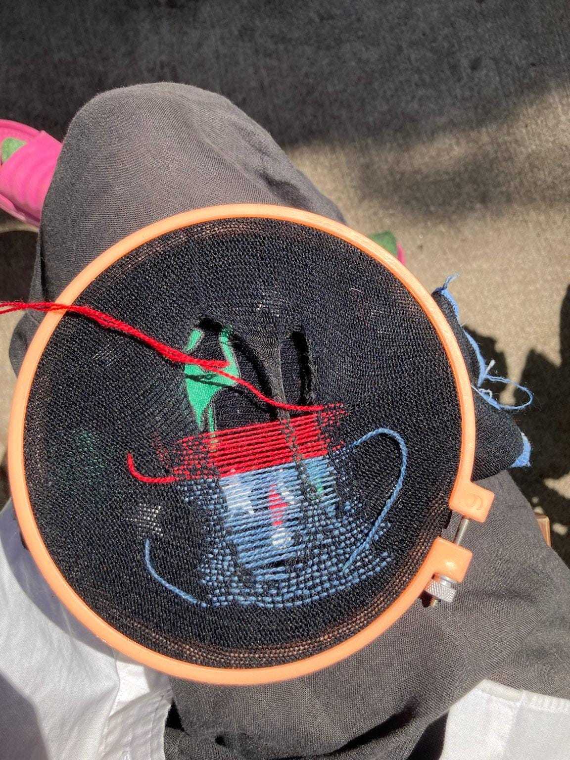 An orange embroidery hoop sits on Khanh's leg, with a sock on it with 2 big holes. Red and blue yarn are threaded through the sock horizontally