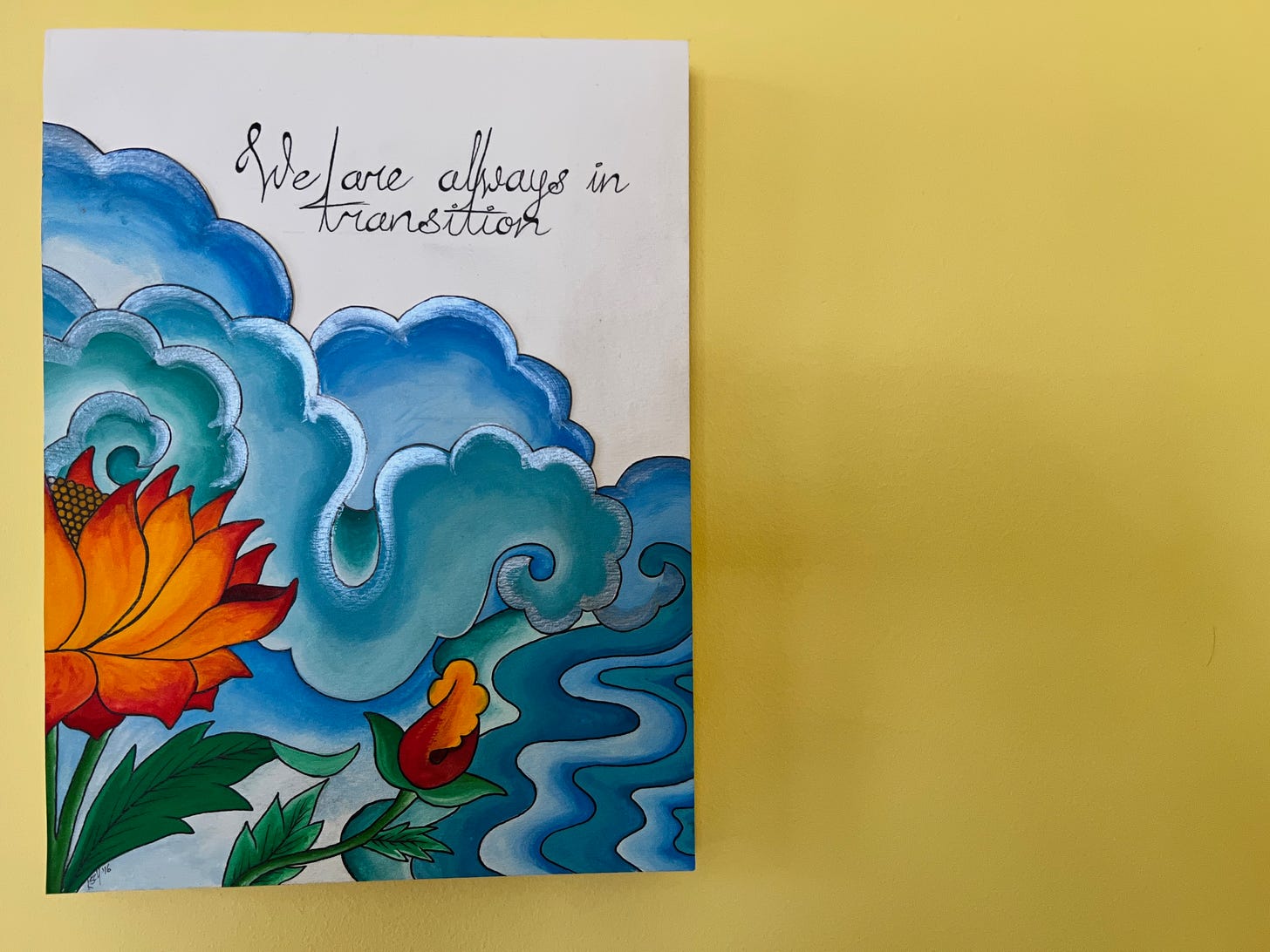 Gouache and ink art piece hanging on a yellow wall. The art shows flowing clouds and water with a lotus flower in the foreground. In black text in the upper right corner it reads: We are always in transition. 