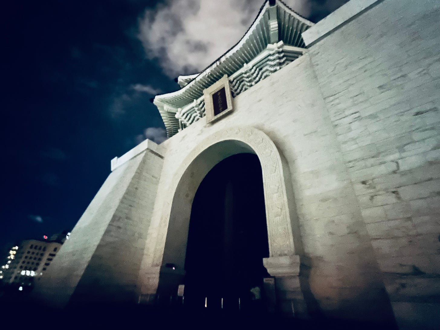A tall, smooth white wall framing an archway into a pitch black entryway. The sharply upturned camera angle also reveals the underside of eaves decorated to look like the interlocking wooden brackets of a traditional Chinese palace.