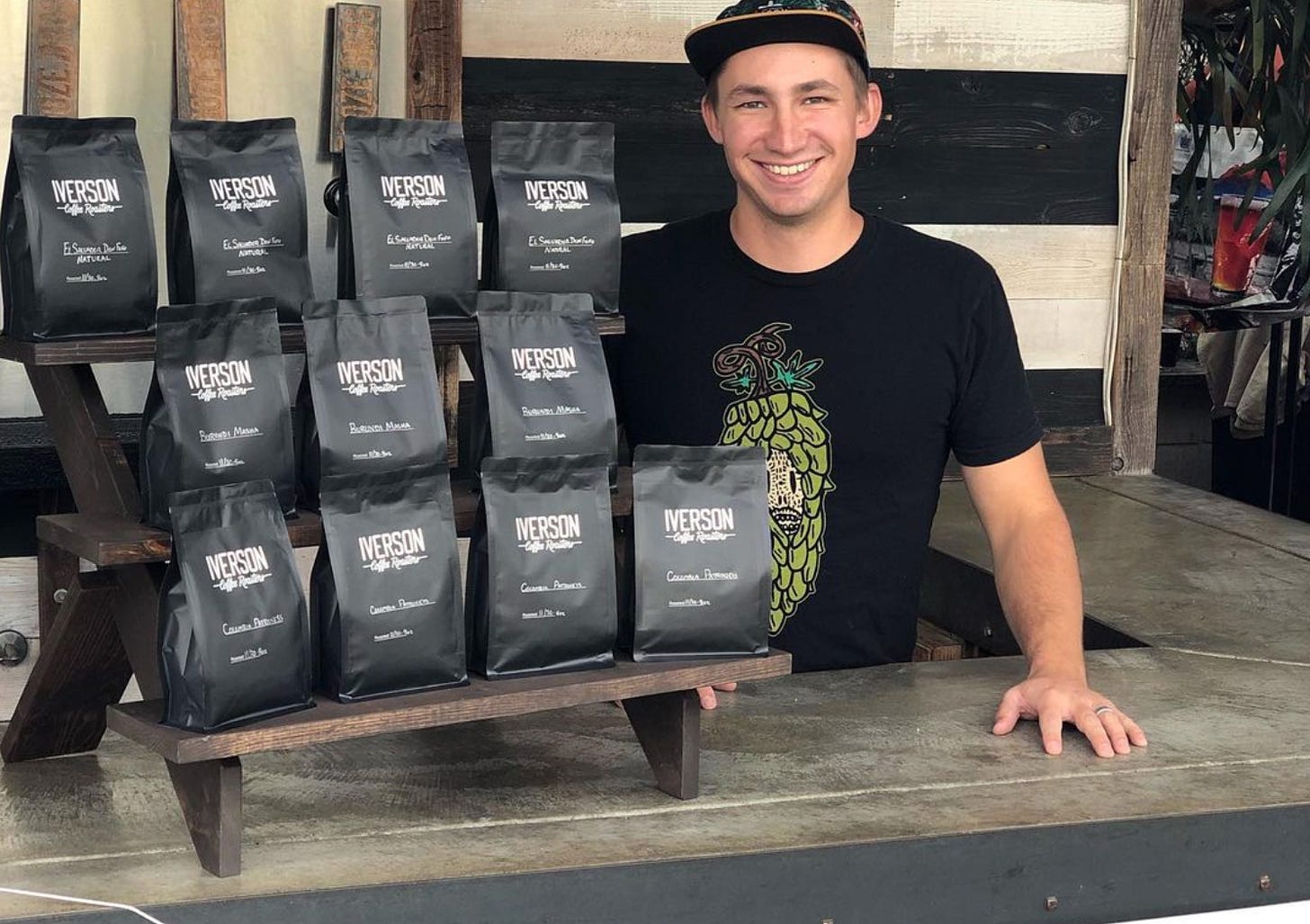 On the left three tiered rows of black coffee bags labeled "Iverson Coffee Roasters," and to the left, just behind the coffee bags is the roaster Danny Iverson.