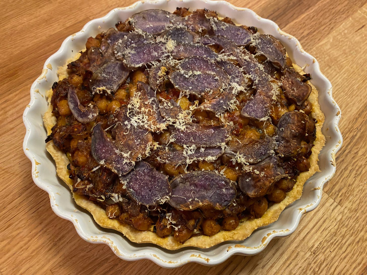 A round tart in a white fluted ceramic tart pan sits on wooden counter. The top of the tart is covered in thinly sliced purple potatoes and grated Parmesan. The chickpea filling is visible underneath the potato layer.