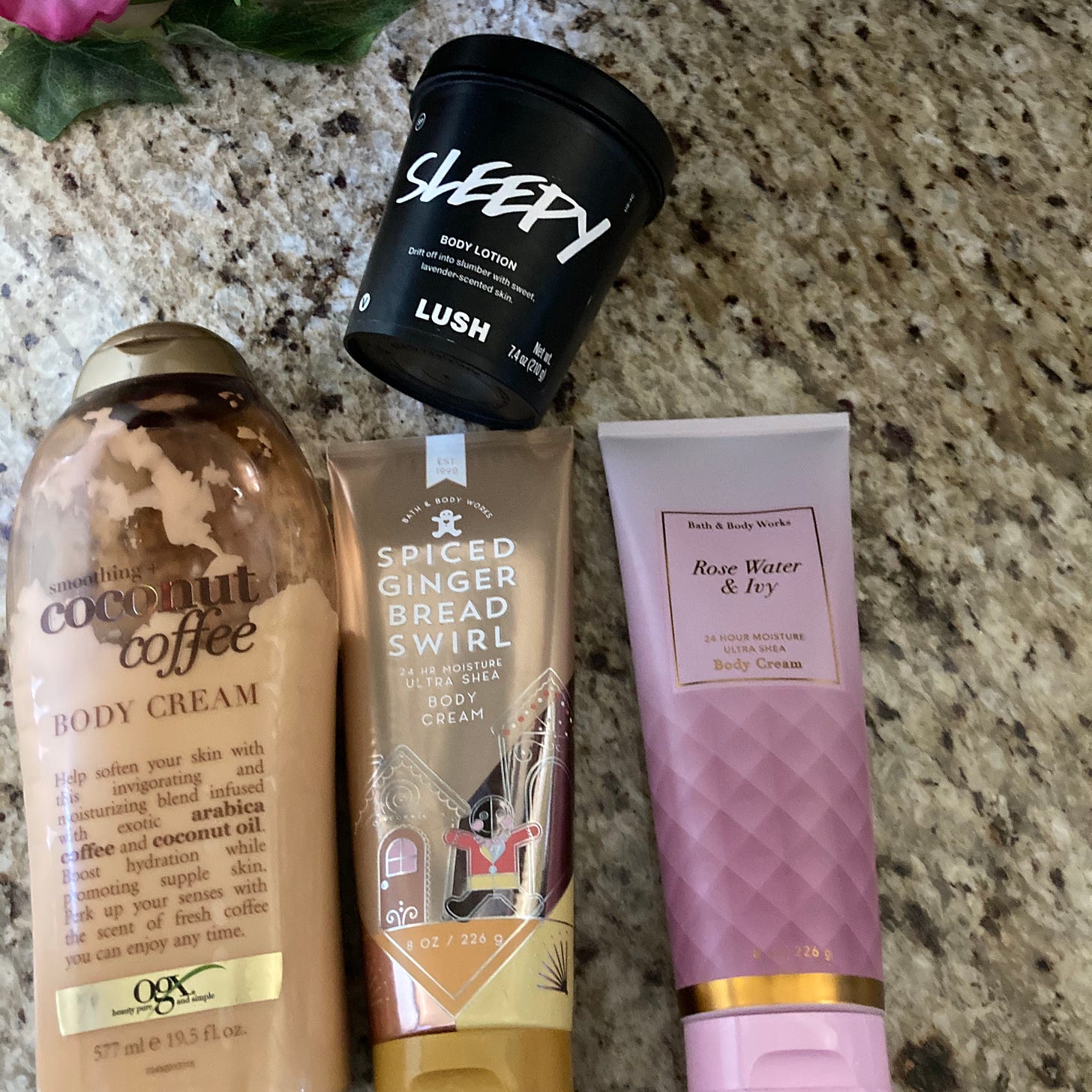Visual description: A brown OGX coconut coffee lotion bottle, a shiny copper Bath and Body Works spiced gingerbread swirl lotion tube decorated with gingerbread men, a pink gradient Bath and Body Works rose water and ivy lotion tube, and a black Lush pot of Sleepy lotion all rest close together on a gray and brown granite countertop.