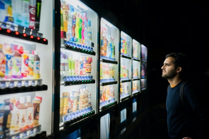 A man staring at a whole bunch of drink options across several vending machines with a concerned/frustrated look on his face like he can’t choose.