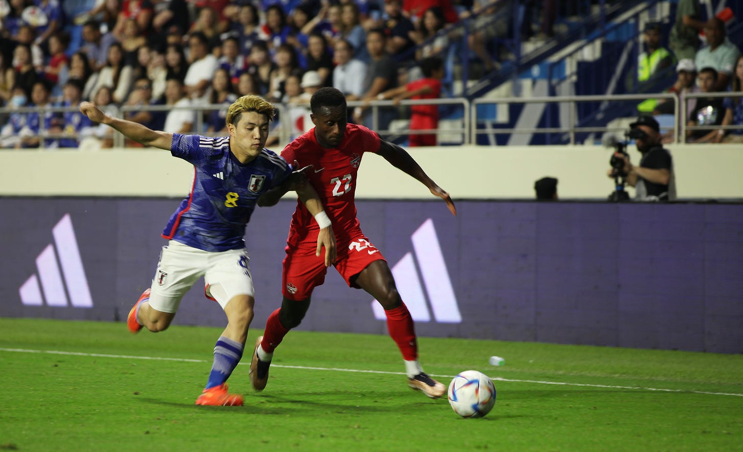 Japanese footballer Ritsu Doan chases Canadian footballer Richie Laryea on the pitch, with the ball ahead of them and the crowd in the background.