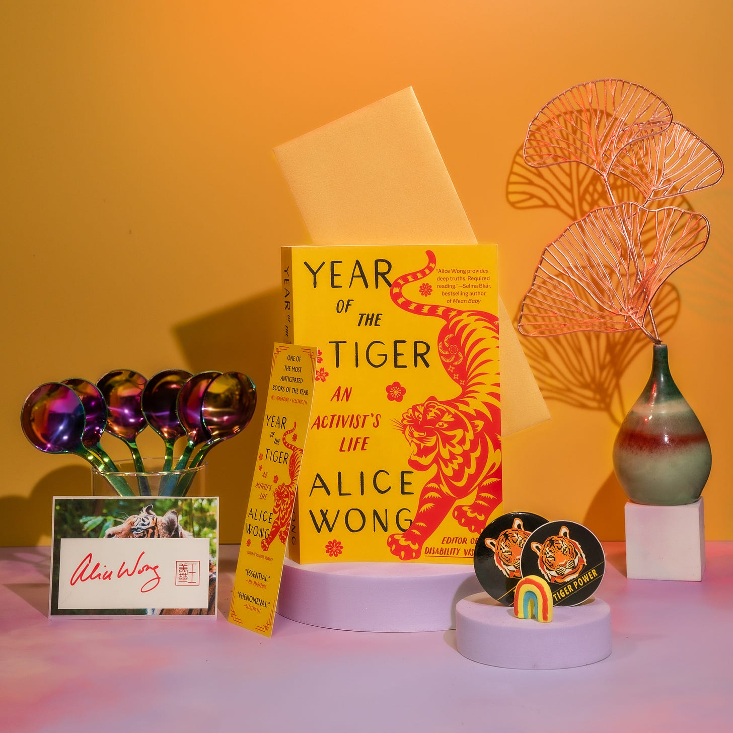 Photo with a tangerine background with a pink border at the bottom with a copy of Year of the Tiger paperback in the center. The left in a glass cup is a bunch of rainbow dipper spoons from @umeshiso_ with a signed tiger bookplate by Alice Wong in front of the cup. A tiger bookmark positioned to the left of the book. On the right is a multicolored pear-shaped vase holding gold ginkgo leaf decorations. In the front is one round tiger sticker and one round tiger button with a small rainbow decor standing upright. Photo credit: @ziru.mo (on IG)