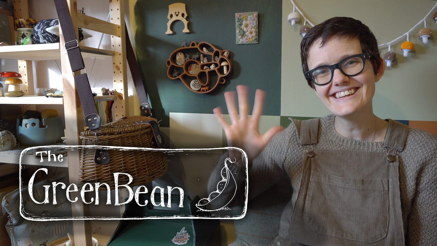 Image description: Title card for a Green Bean Podcast. Katie sits waving at the camera, in front of a wall pantined in squares of differing shades of green. The wall is adorned with a crochet mushroom garland, a ceramic curiosity display, a cello bridge and some floral embroidery. A wicker backet bag hangs from th shelves to the left.