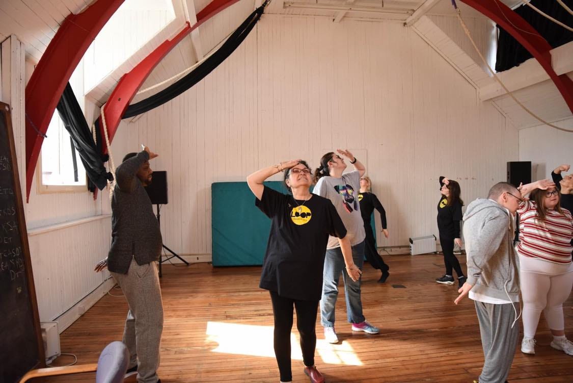 Loop Theatre, a mix of genders and ethnicities, are performing in a space with a wooden floor, white walls and red beams. They all have their right hand up and are resting the right hand against their foreheads.
