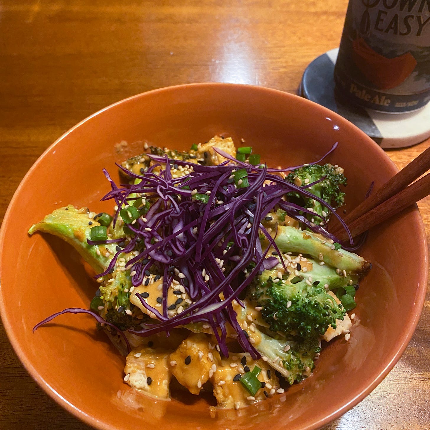 In an orange bowl with chopsticks at the edge are rice noodles, tofu, and broccoli tossed in peanut sauce. On top is a little pile of thinly sliced red cabbage, and a sprinkling of toasted white and black sesame seeds.