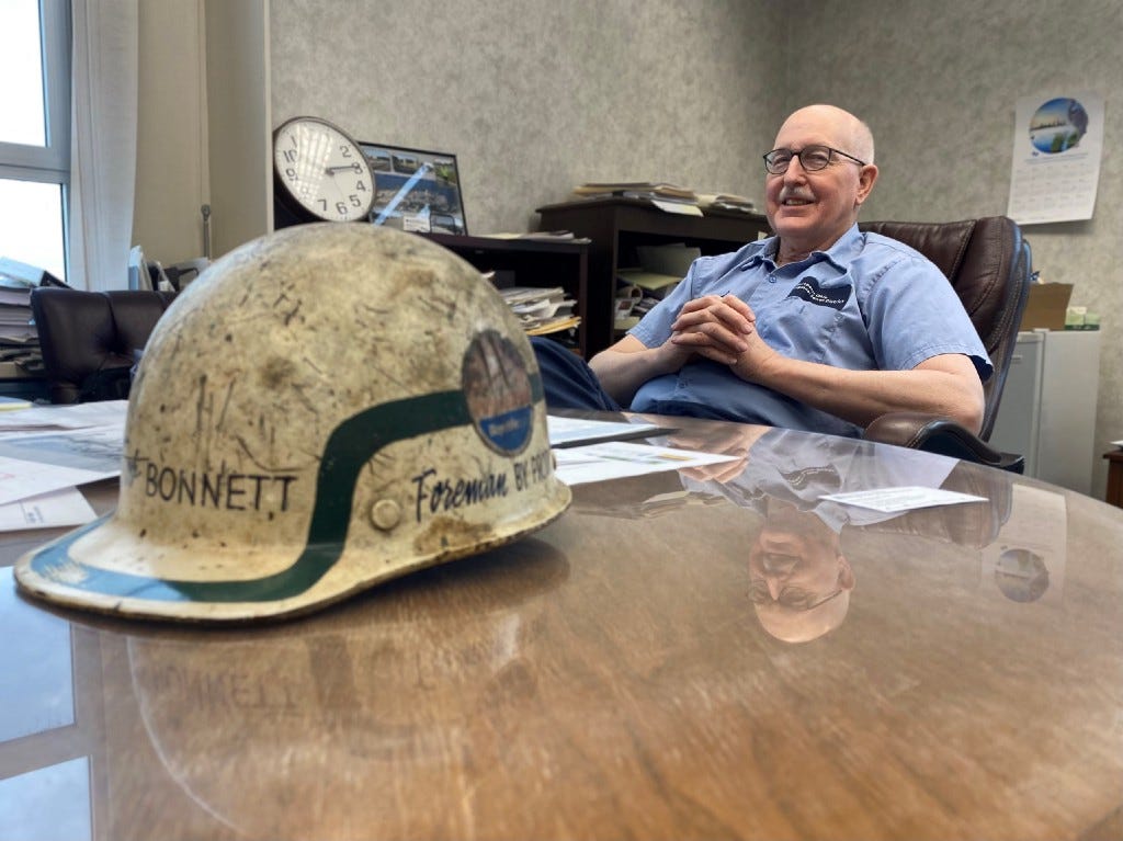 Bob Bonnett leans back in his soft office chair, hands folded across his chest, smiling as he recounts stories of his early days at the Sewer District. his reflection is seen in the gloss of the office conference table, and in the foreground is a weathered and worn hard hat, the same one he wore as a foreman in the steel mills of the 1970s.