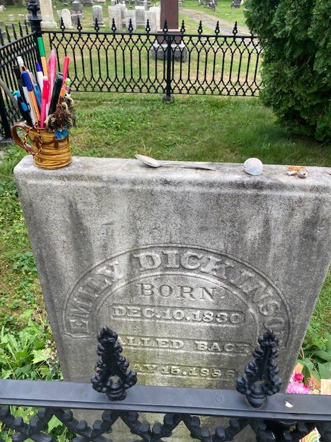 Image of a tombstone with pens in a cup and a spoon resting on top. the tombstone reads Emily Dickinson Born Dec 10 1830. 