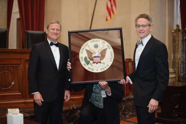 Two men in tuxedos smile as they stand on either side of a framed Supreme Court seal, which is being held up by a person standing behind the frame whose face is not visible. In the background is the bench where the justices sit.