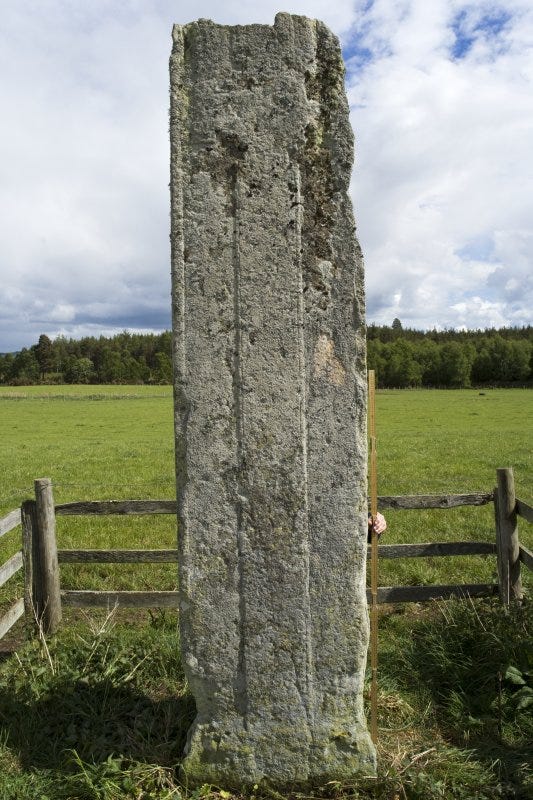 The Altyre Cross, photographed standing in a field on the Altyre Estate. It's a tall, grey sandstone slab, very worn, with a thin cross just discernible.