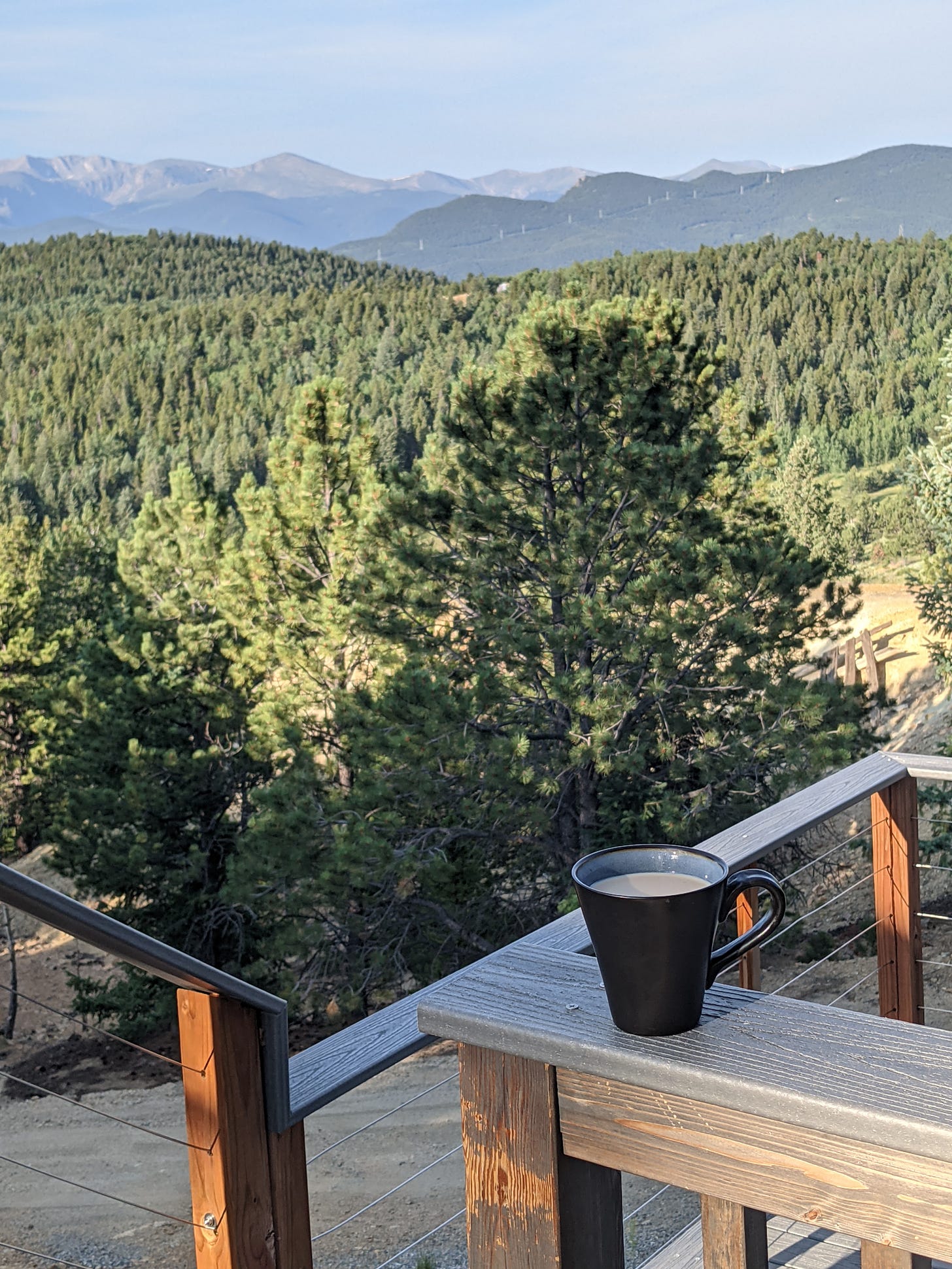 A black coffee mug full of milky coffee sits on a blond wooden porch railing. A wooden deck with fencing continues down stairs, before a view of green pine trees and in the near background, rows of brown and blue mountains below a pale blue, morning sky.