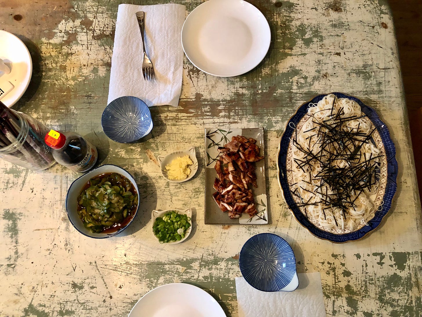 Overhead shot of a table with dishes of cold somen noodles, grilled chicken, and vinegared cucumber slices. There's also a bottle of store-bought dipping sauce for the noodles.