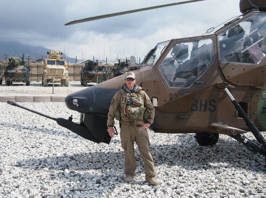 Brian R Price in Afghanistan, standing in combat gear in front of a number of French and American combat vehicles, and a French attack helicopter.
