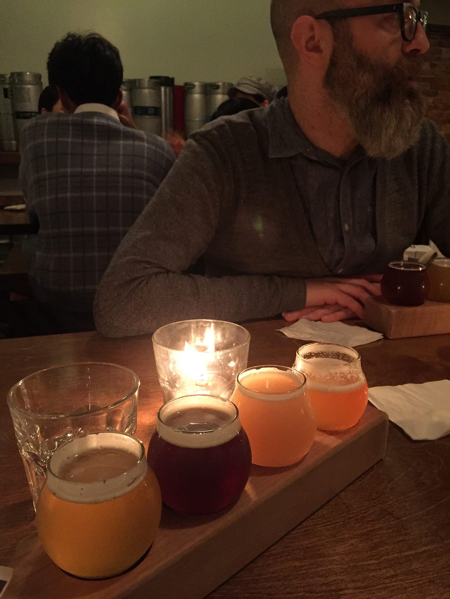 a flight of 4 beers in varying colours sits at an angle in the foreground, lit by a candle in a glass behind it. In the background, Iain, a bearded man in a light blue shirt, sits with his arms on the table in front of his own flight of beer, looking slightly to his left.