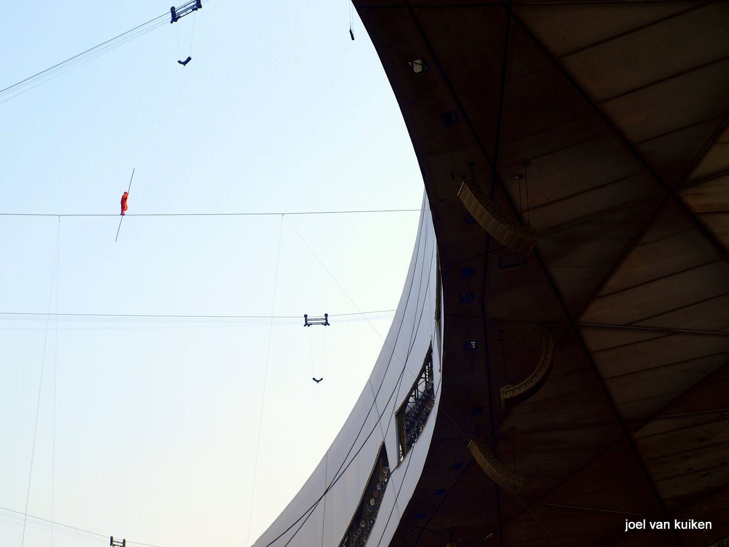 Man in red suit tightrope walking across a stadium. 