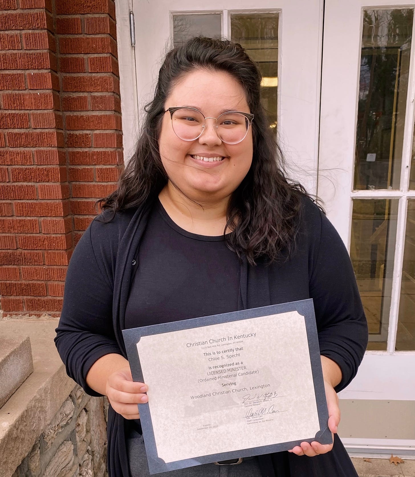 Chloe, a white femme person with brown hair and glasses, is pictured from the waist up. She is wearing all black clothes and holding her certificate of licensure as a pastor. 