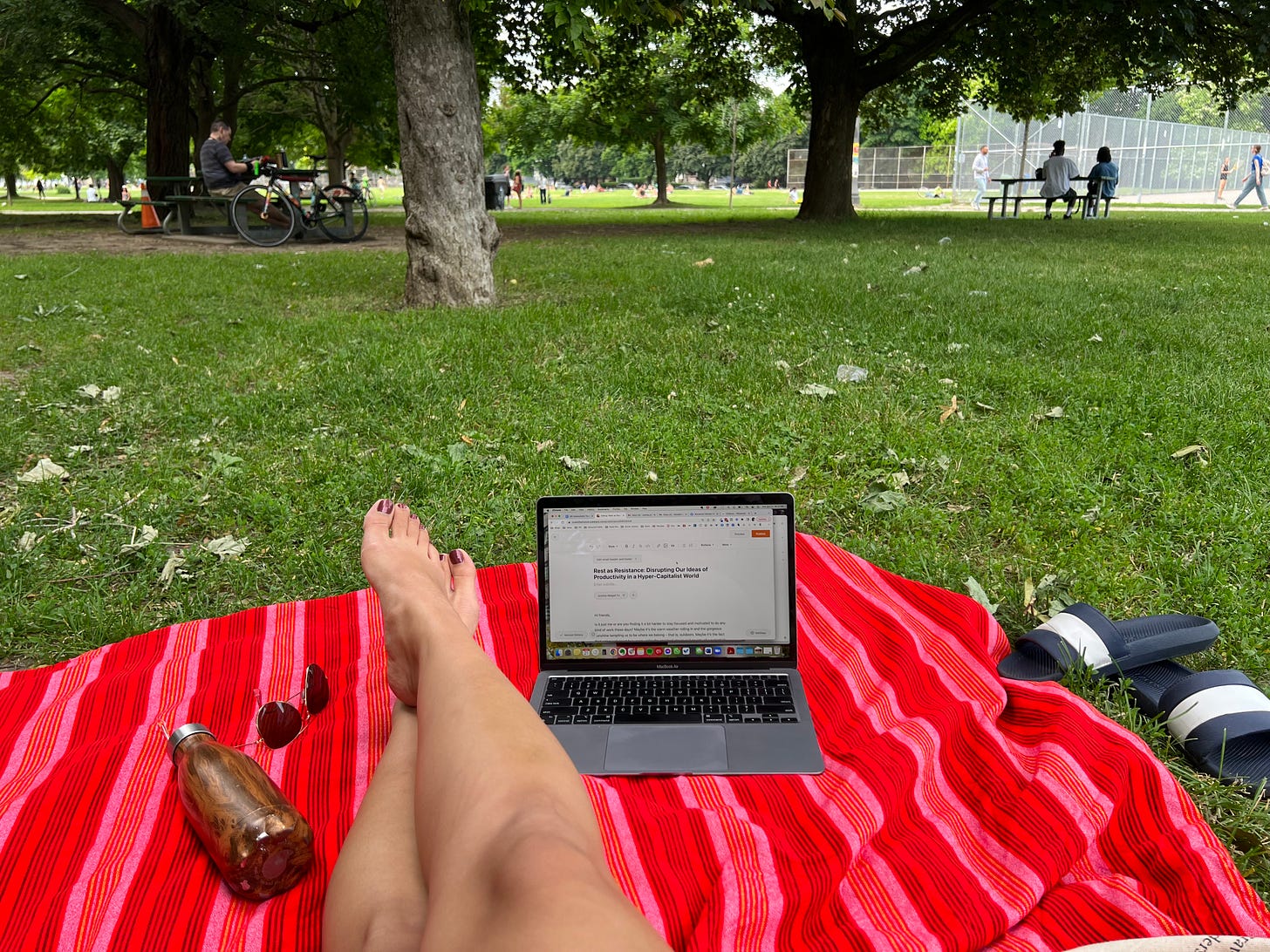 Woman's legs and a laptop on top of a red picnic blanket with a park in the background.
