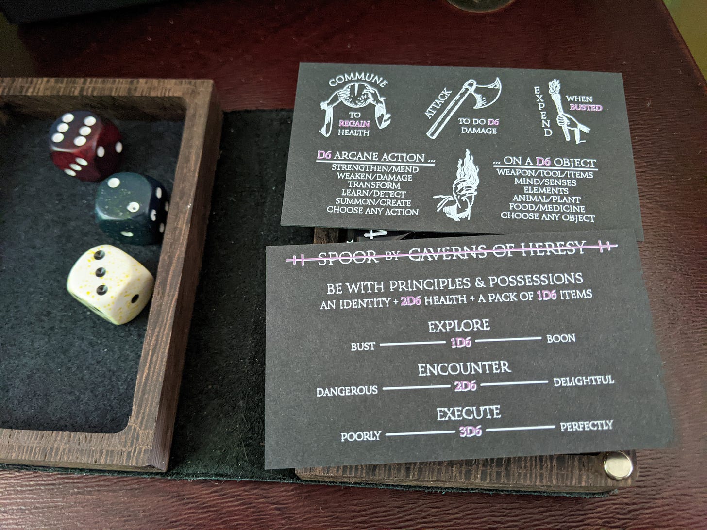 A picture of a dice tray and the front and back of Spoor - a business card rpg