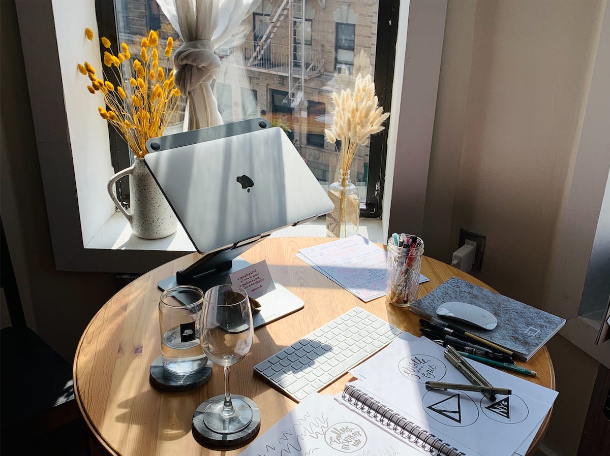 Pictured: Rays of light piecing through a window onto a circular wooden table, its surface scattered with working utensils including a laptop, keyboard, mouse, water, wine glass, notebook, and illustration pens.