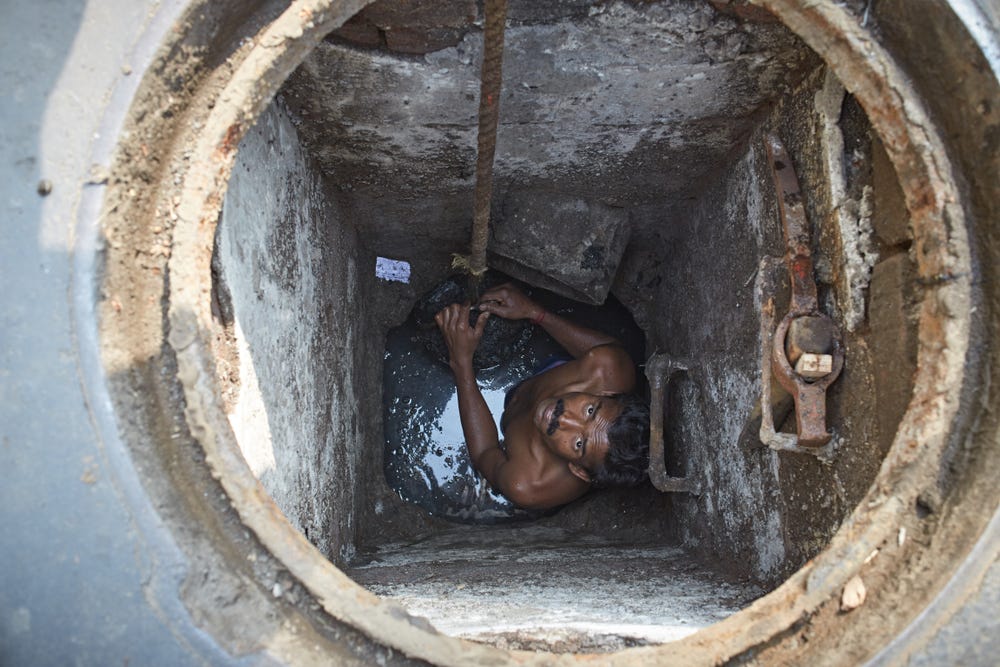 A man with a moustache and dark skin looks up from a sewer hole. He is standing in liquid and holding on to a rope.