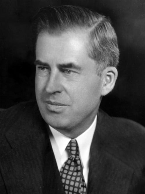 Black-and-white image of the head and shoulders of man about fifty with upswept hair, wearing a gray suit and a dark tie