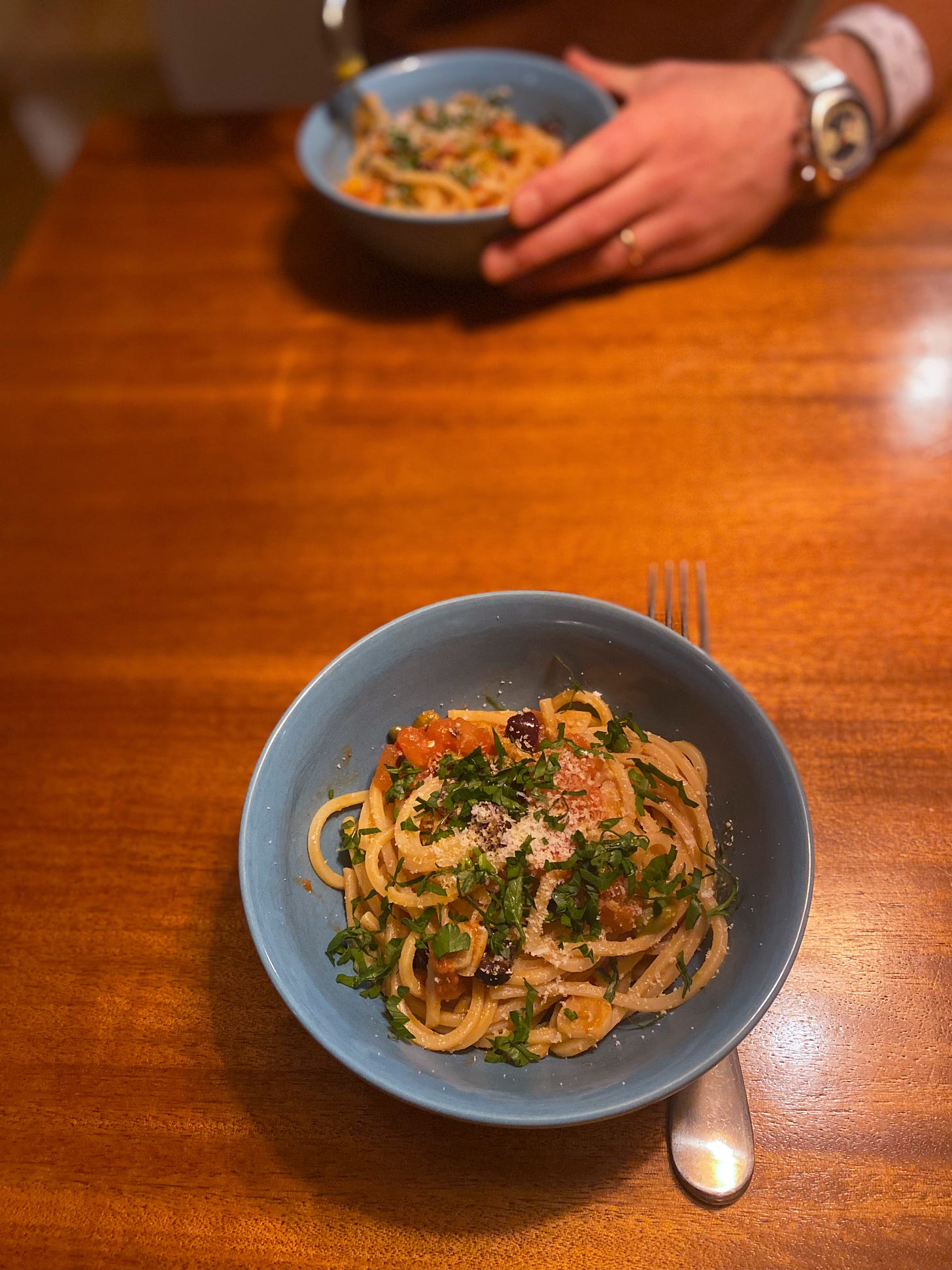 Two blue bowls of pasta across from each other on the table. Each is dusted with parmesan and a sprinkling of chopped parsley. Jeff's hand is curled around his bowl on the far side of the table, fork hovering above his food.