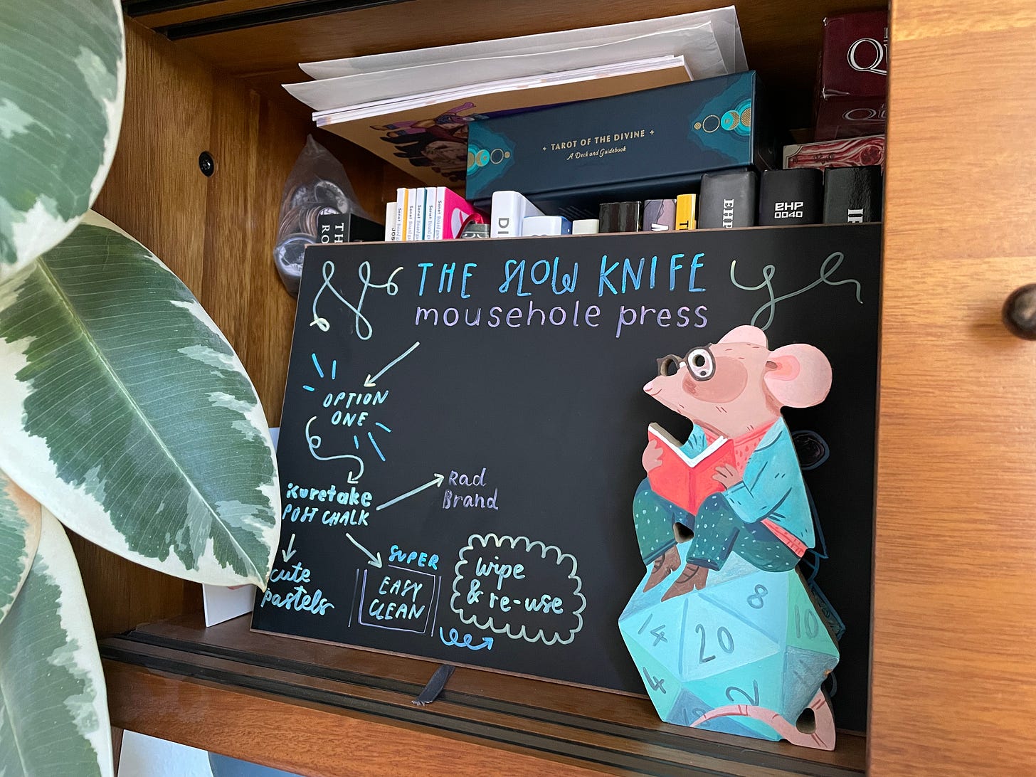 A decorated chalk board that reads 'The Slow Knife, Mousehole Press'. It has a load of arrows and boxes in different chalk colours. Behind, there are some books stacked up and in front there is a wooden standee of the Mousehole Press logo.