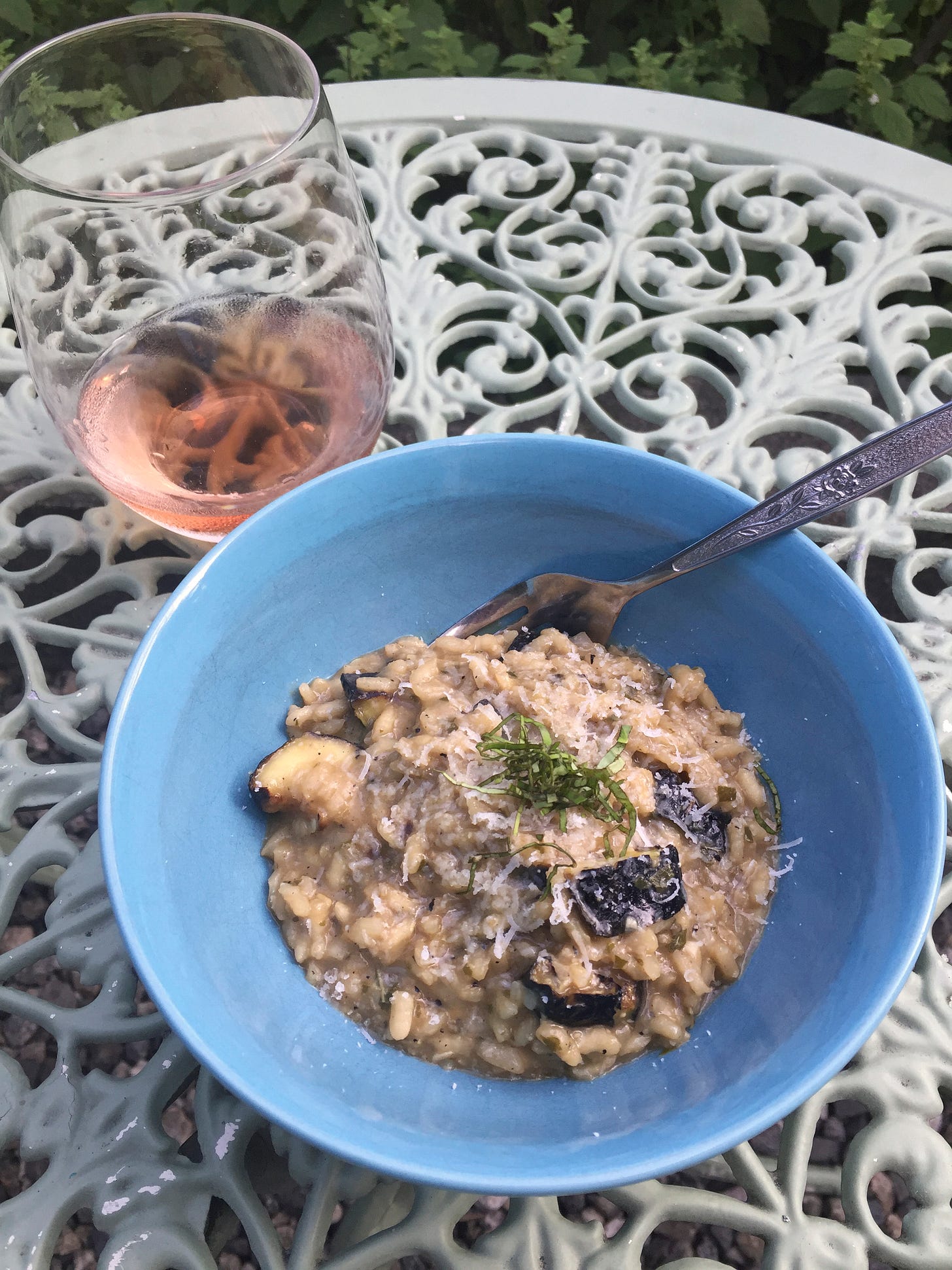 A blue bowl of risotto with pieces of grilled zucchini throughout, and grated pecorino and thinly sliced basil are sprinkled over the top. A fork sticks out at the back of the bowl, and a glass of rosé sits on the table beside it.
