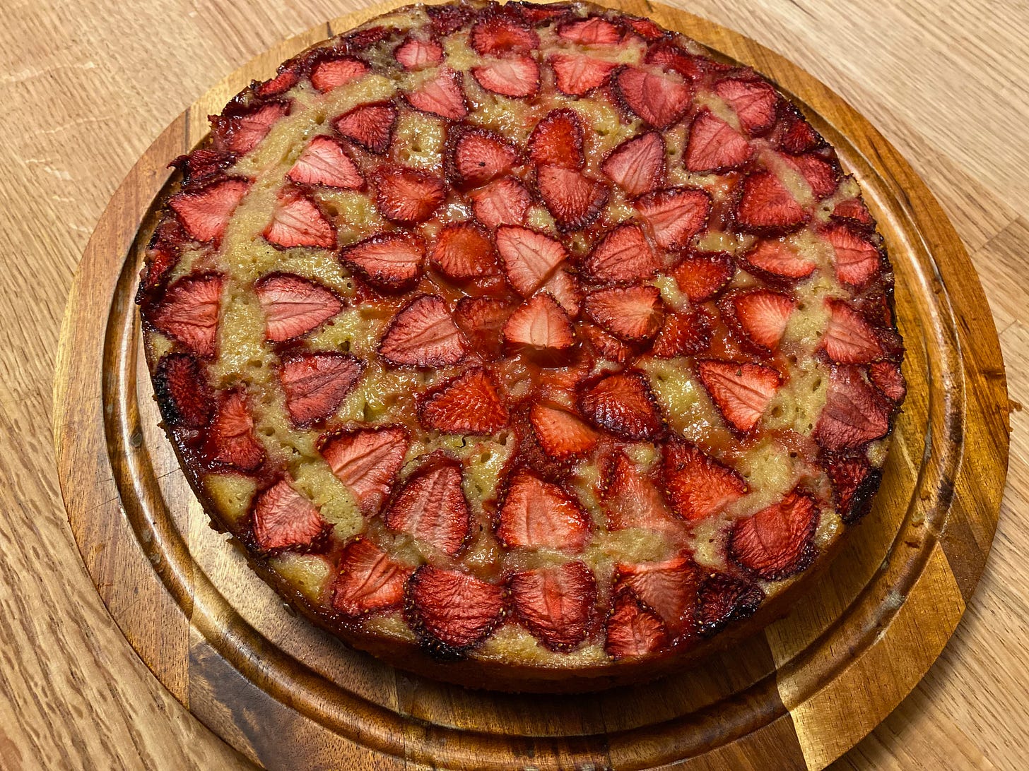 A round cake covered in sliced strawberries arranged in circles sits on a wooden counter.