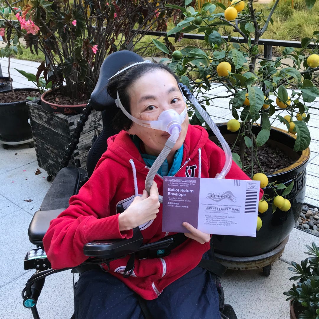Asian American disabled woman in a wheelchair wearing a red hoodie with a mask over her nose attached to a gray tube. She is sitting in an outdoor patio in front of a potted lemon tree. She is holding an absentee ballot.