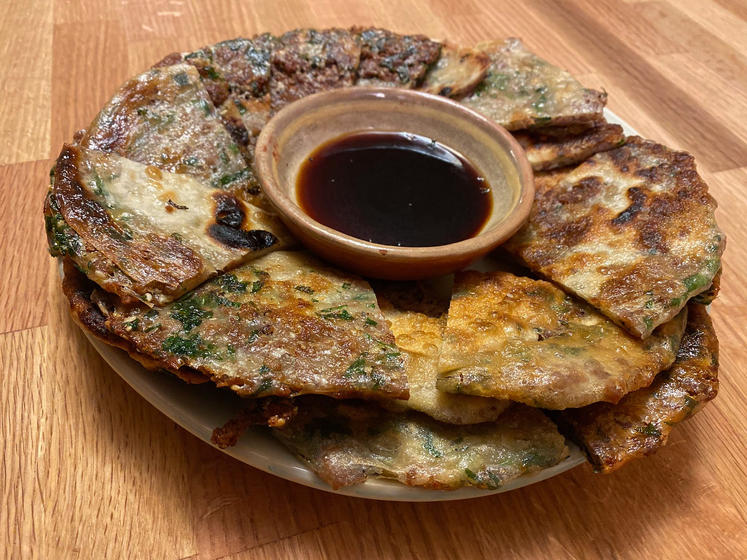 An overflowing plate of golden-brown pancakes, cut into triangles, sits on a wooden counter. There’s a small bowl of soy sauce in the middle of the plate.