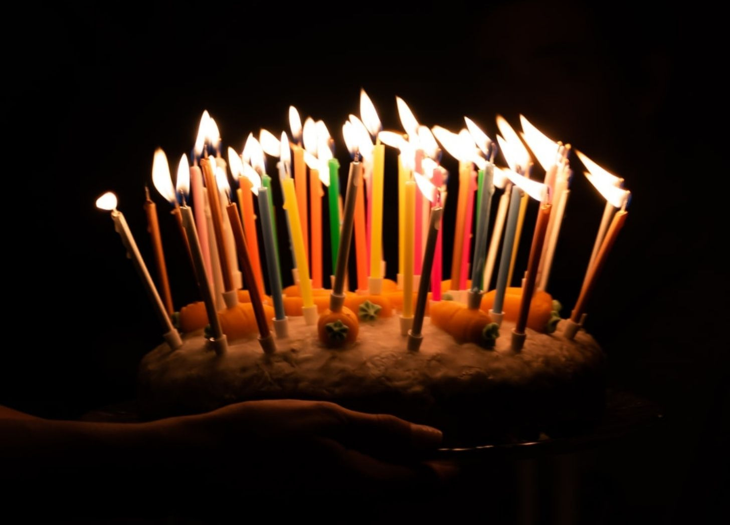 Foto de mãos segurando bolo de aniversário com muitas velas acesas em um ambiente totalmente escuro.