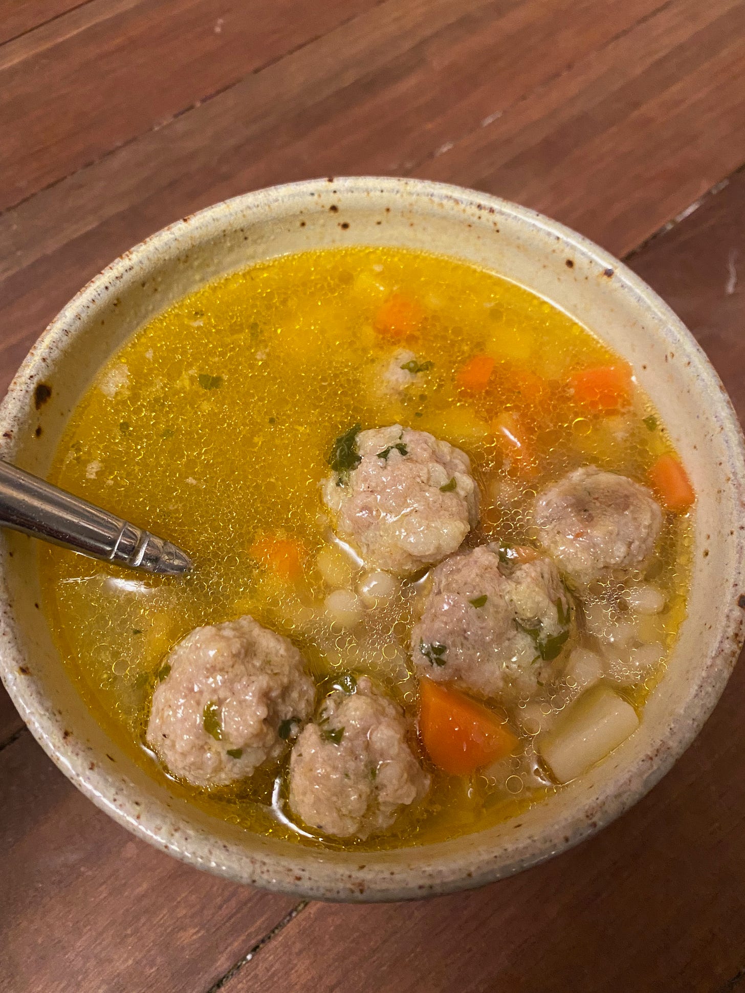 A ceramic bowl filled with deep golden chicken broth. Several small meatballs float on top, along with small pieces of carrot, and pearl couscous.