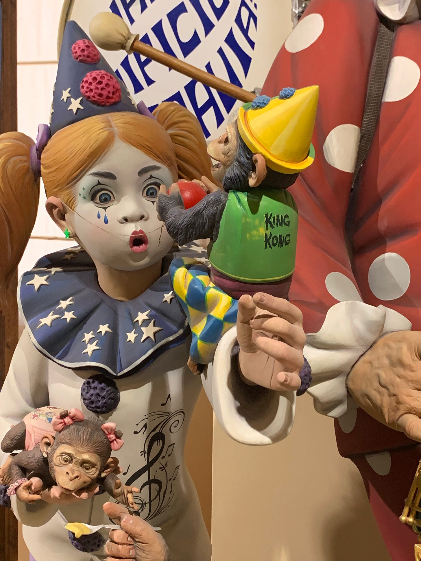 A ninot of a little girl dressed as a clown holding a tiny chimpanzee who is also dressed like a clown and is about to snap her red clown nose at her face. His vest says "King Kong" on the back.