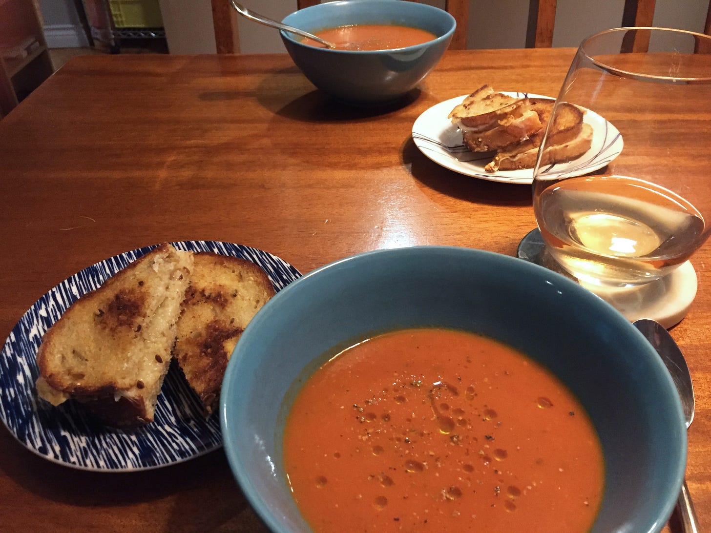Two blue bowls of tomato soup with drizzles of olive oil and bits of thyme. Beside each of them is a small plate with a grilled cheese sandwich, and a glass of wine.