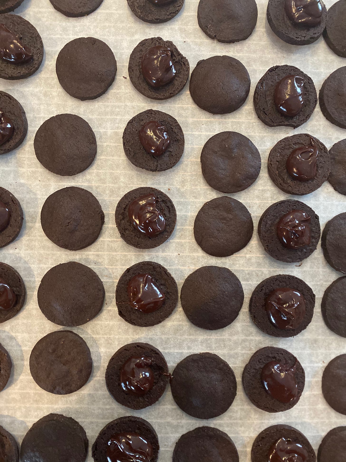 Many chocolate O cookies sit on a sheet of parchment paper waiting to be sandwiched. There are alternating rows of plain round chocolate cookies, and plain round chocolate cookies topped with a dollop of chocolate ganache.
