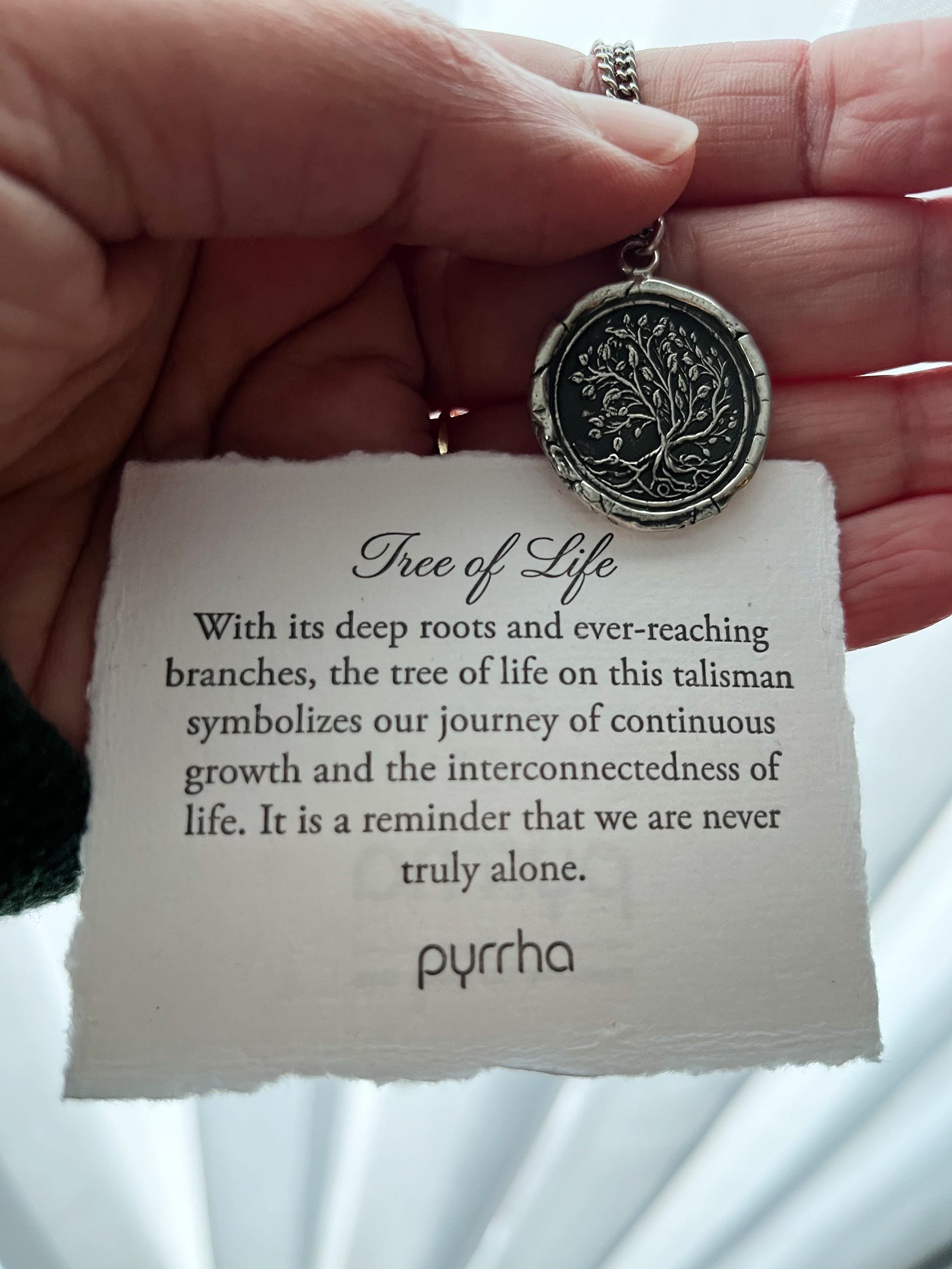 Photo of Olwen’s hand holding a Pyrrha silver pendant with an image of a tree with branches, leaves and roots on it. The white card behind it reads: “Tree of Life. With its deep roots and ever-reaching branches, the tree of life on this talisman symbolizes our journey of continuous growth and interconnectedness of life. It is a reminder that we are never truly alone.”