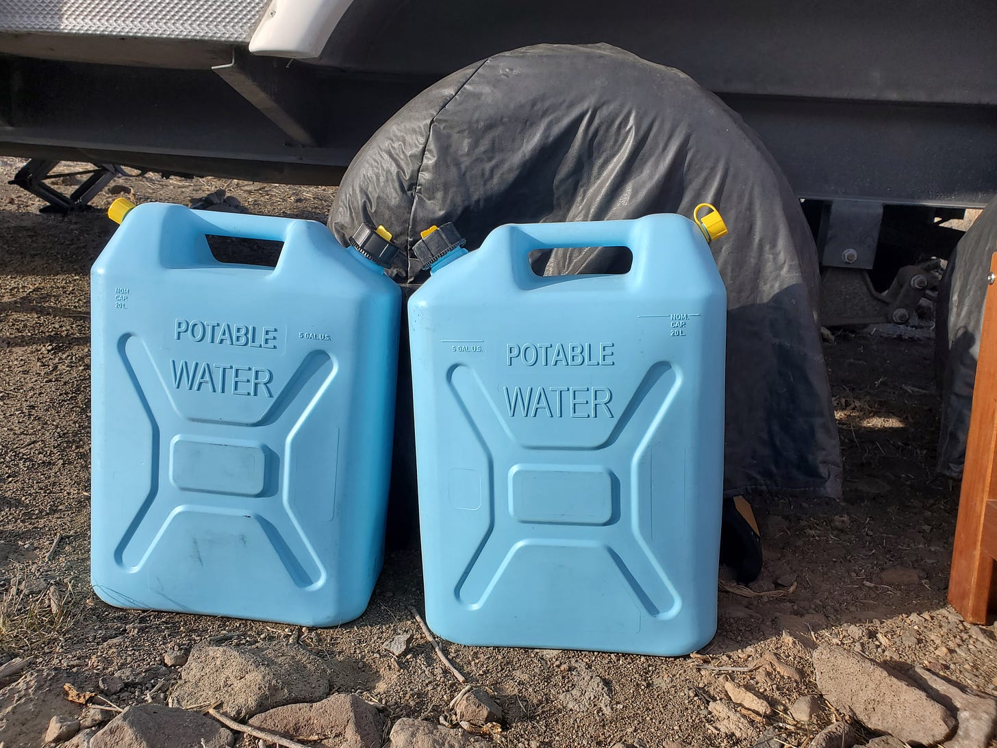 Two blue plastic containers that say Potable Water on the ground in front of a wheel of the travel trailer.