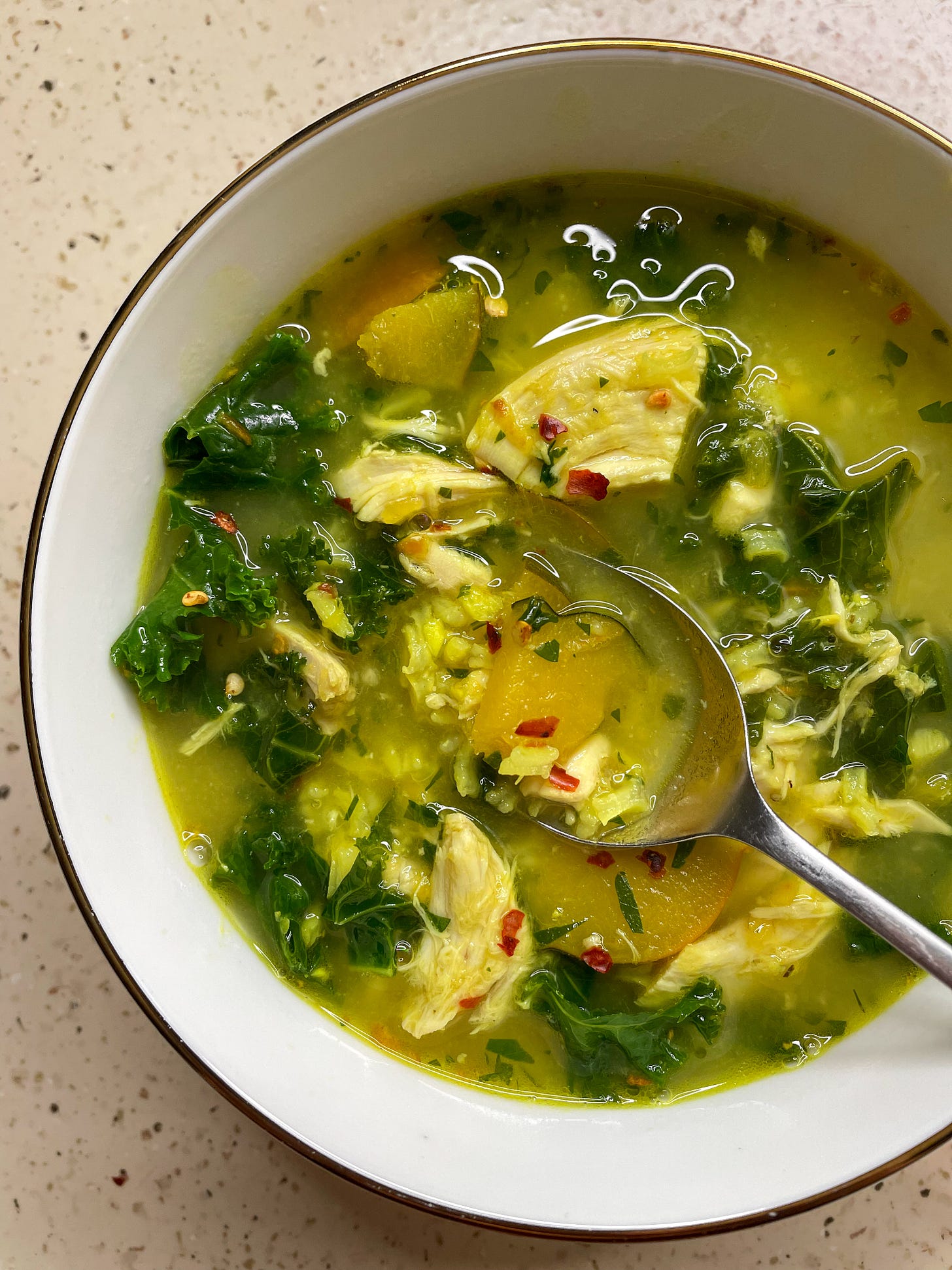 A bowl of fragrant, golden ginger and turmeric chicken soup, sprinkled with red pepper flakes and dotted with fall produce like acorn squash and kale.