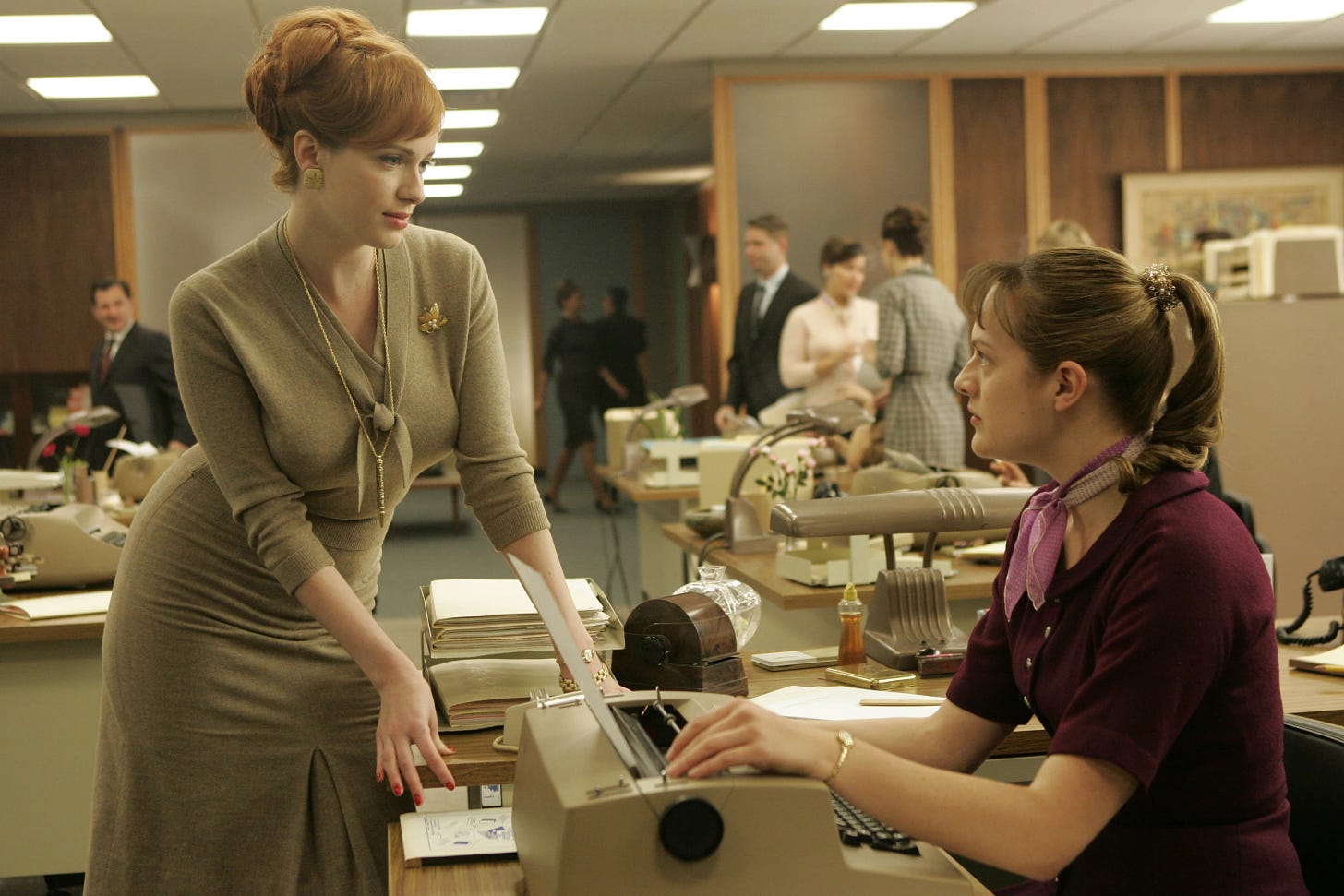 A white 30-something woman in a grey dress hovers over the desk of a 20-something woman in a purple dress, who looks up from her typewriter. They're in a 1960s office.