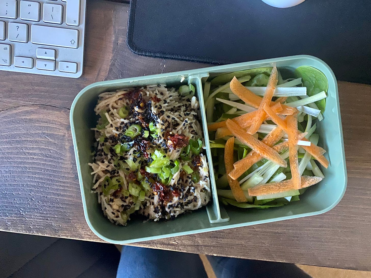 bento box filled with noodles and a salad of cucumber, carrots and spring onions. The green box is placed on a wooden desk, next to a mouse mat and keyboard