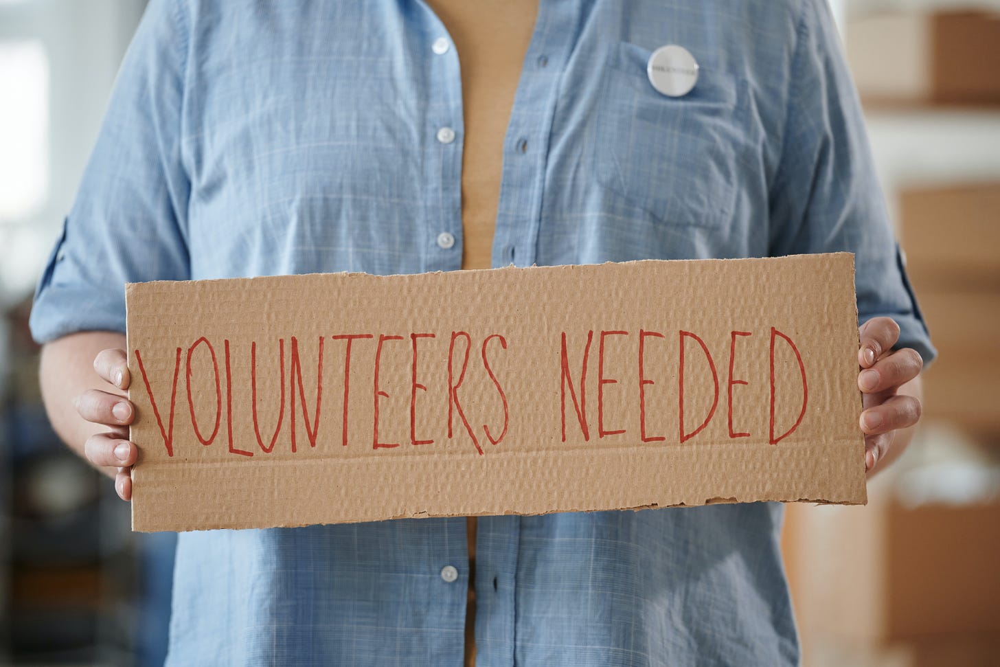 A person wearing a blue shirt holding a sign that says "Volunteers Needed."