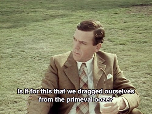 still image from the TV show Jeeves & Wooster showing Hugh Laurie's character Bertie Wooster. He is seated and holding a drink, wearing a tan suit, saying "Is it for this that we dragged ourselves from the primeval ooze?"