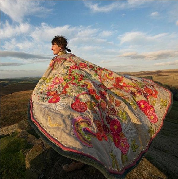 A woman stands on a hill wearing a giant flowing cape made of pale material and embroidered with luciously coloured flowers